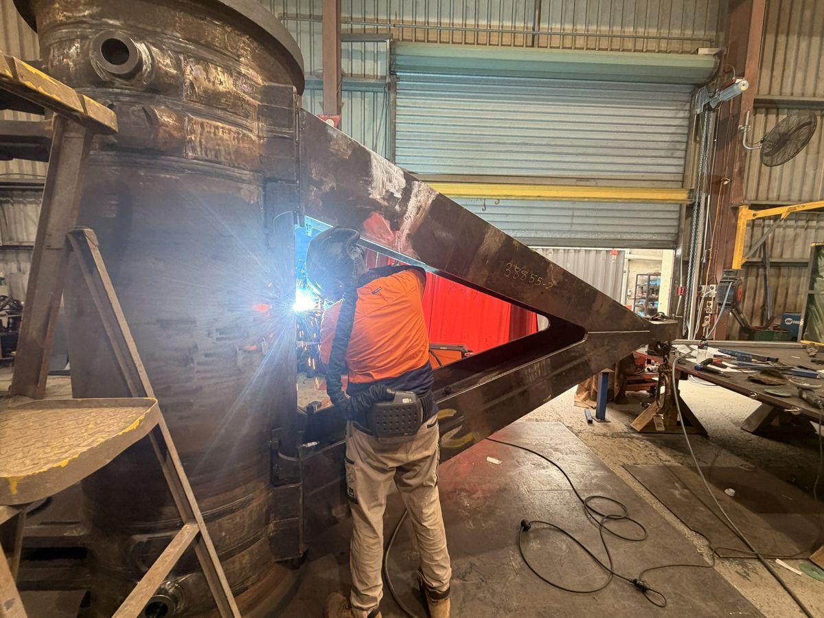 A welder in orange hi-vis welding a heavy axle fabrication in the workshop