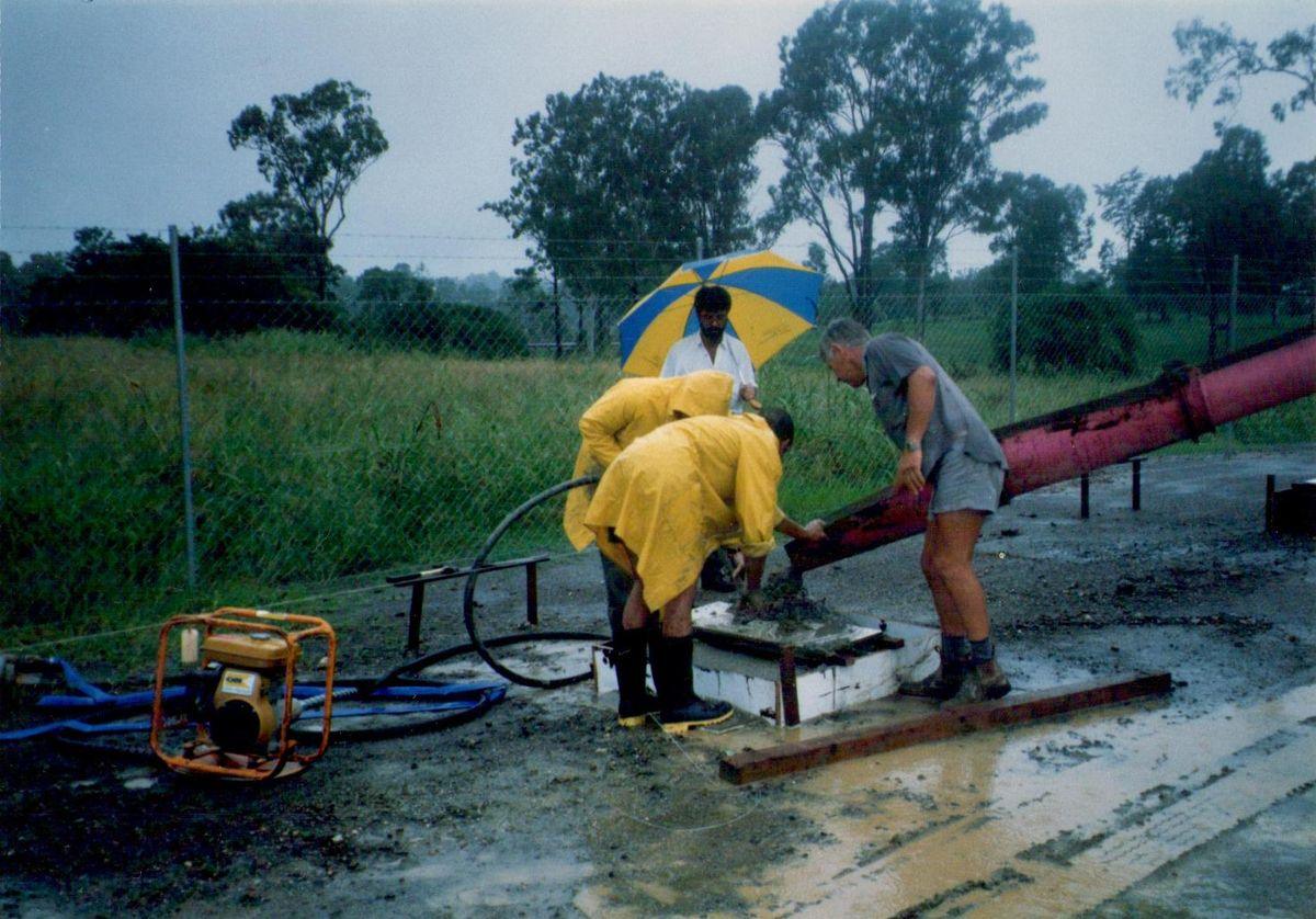 Workers in wet-weather gear pouring concrete foundations in the rain under a beach umbrella