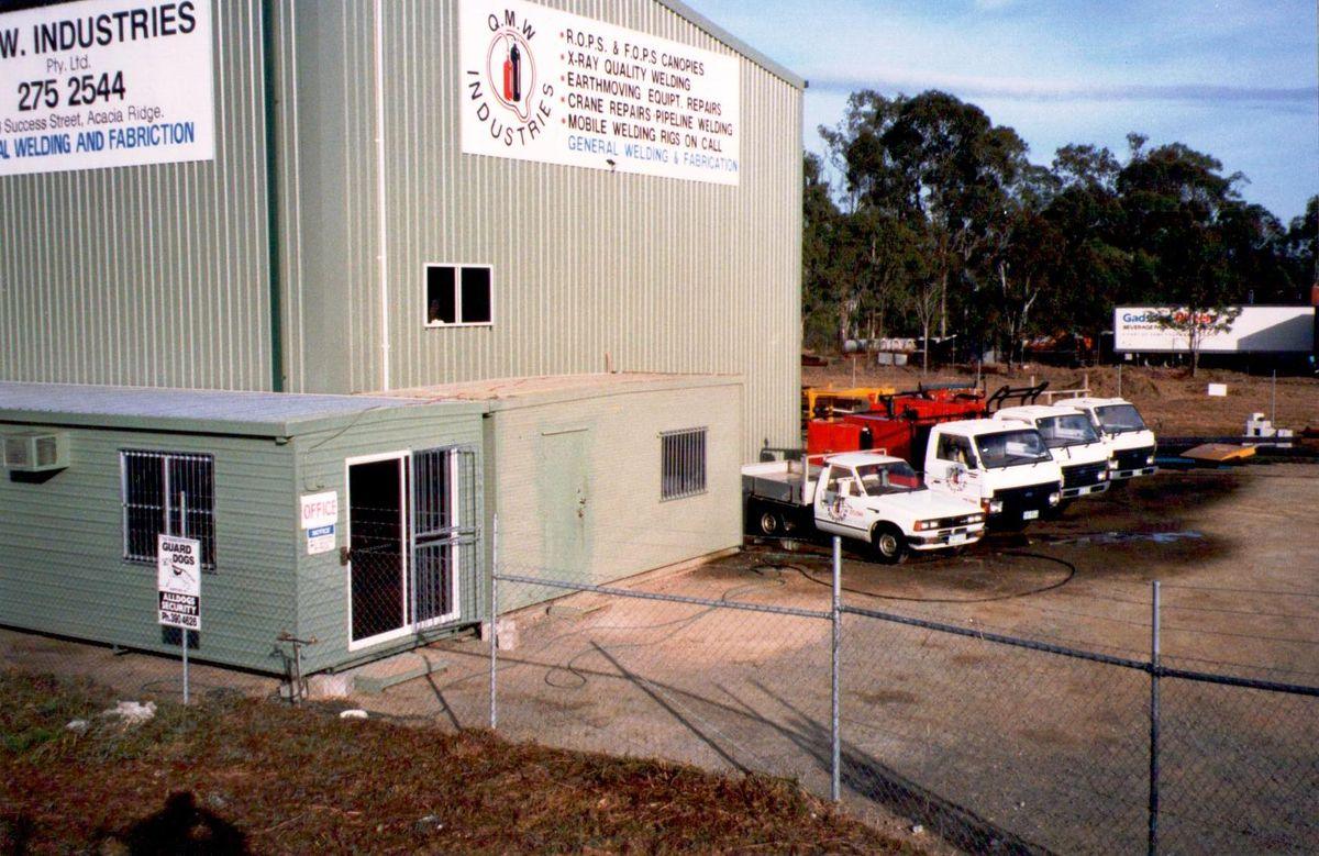 The Acacia Ridge workshop frontage with a row of white service utes and a sign reading Q.M.W. Industries, listing ROPS and FOPS canopies and X-ray quality welding