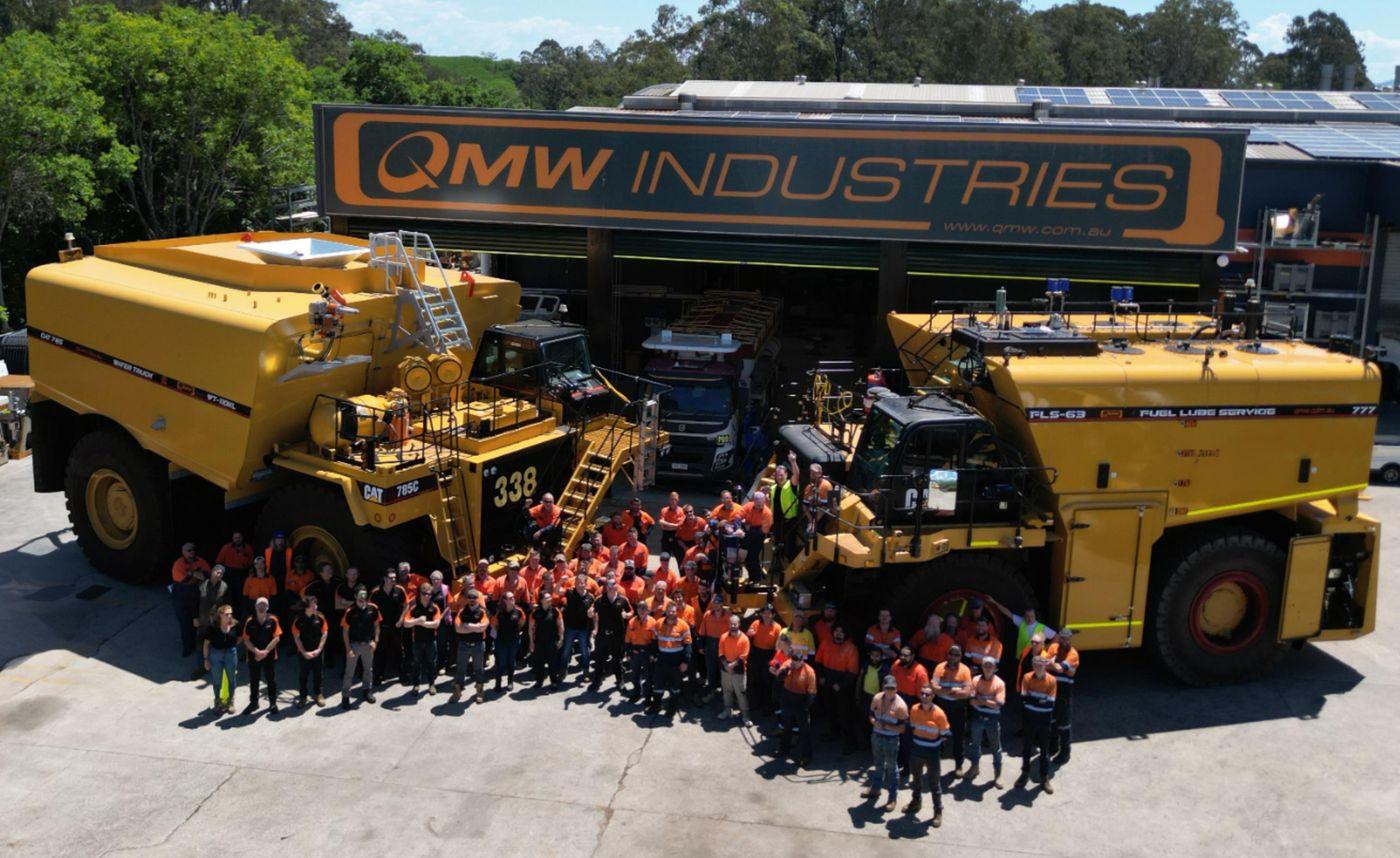 The QMW team in orange and navy workwear gathered in front of the QMW Industries workshop, between a Cat water truck and an FLS-63 fuel-lube service truck