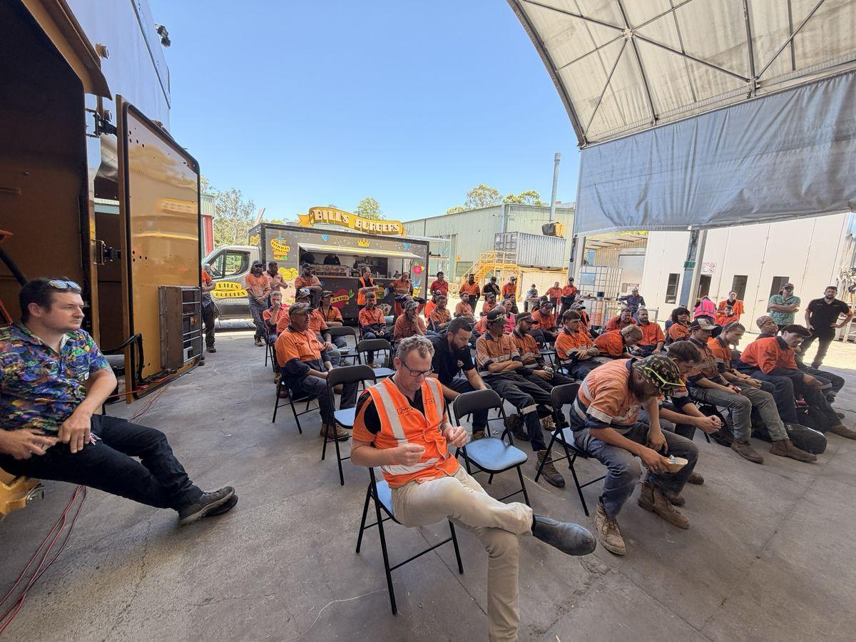The team seated for a toolbox meeting under the front workshop roof