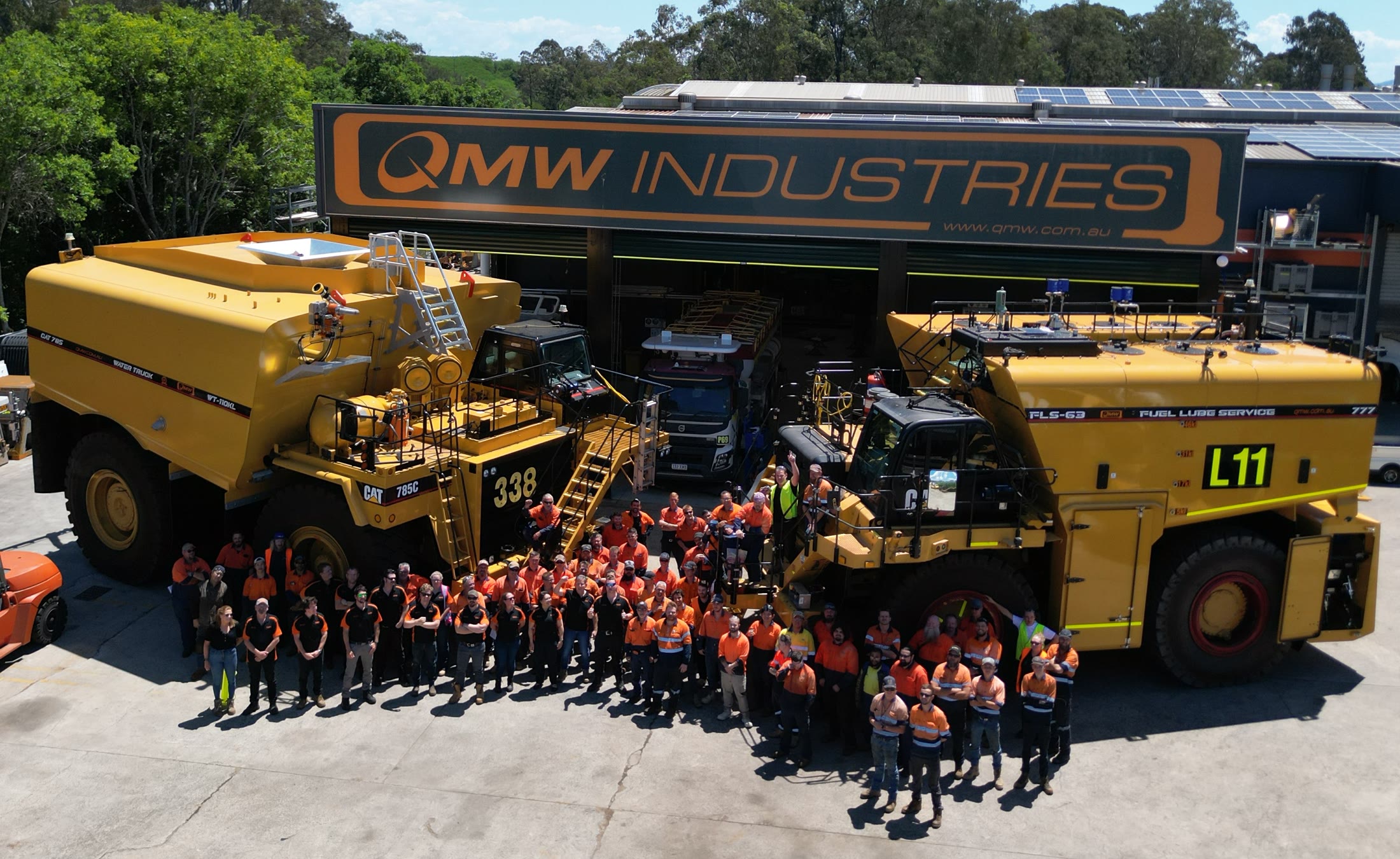 The QMW Industries team outside the Acacia Ridge workshop, framed by a CAT 785C WT-110KL water truck and a CAT 777 FLS-63 fuel and lube service truck